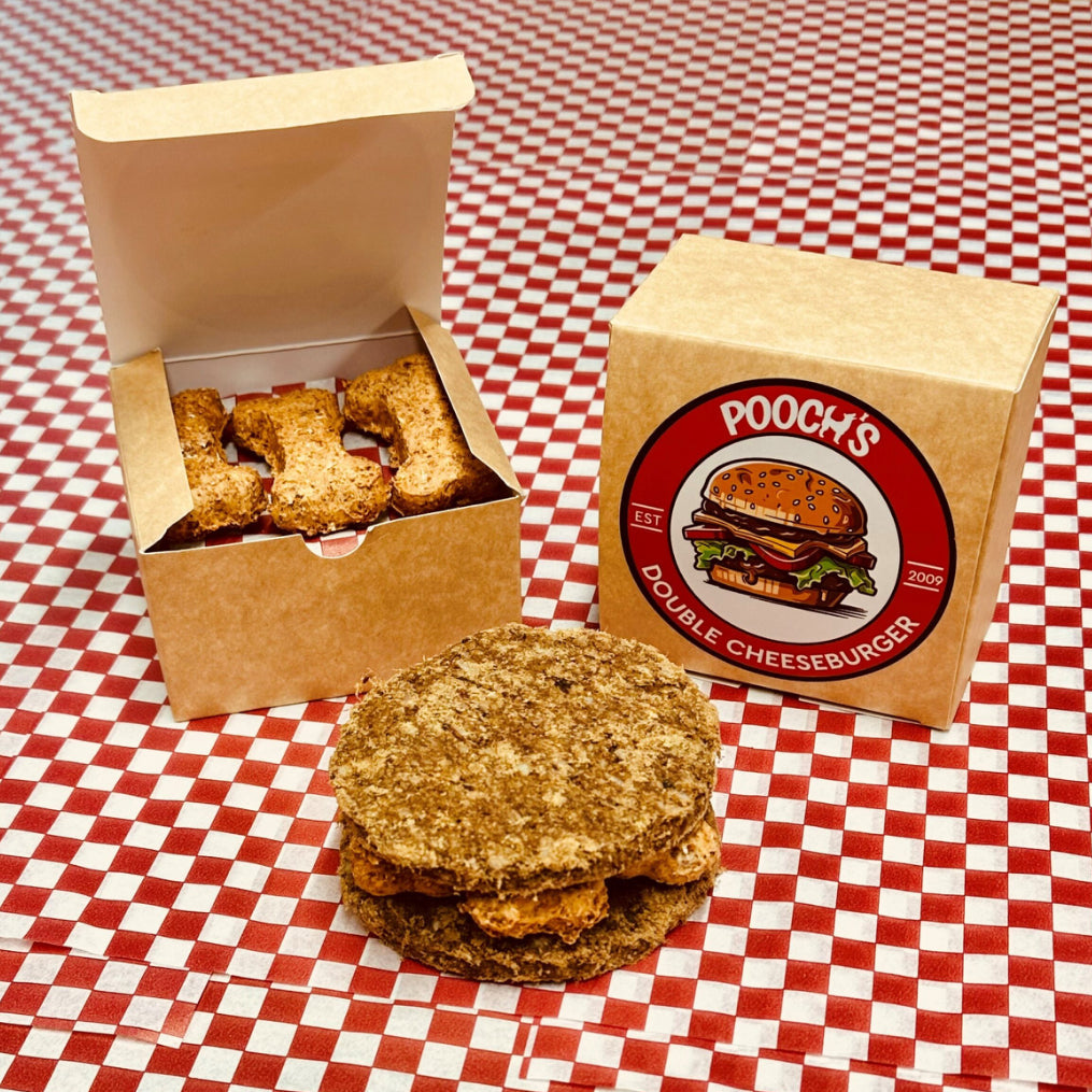 A packaged dog treat resembling a double cheeseburger, with the product visible outside the box, placed on a red and white checkered surface.