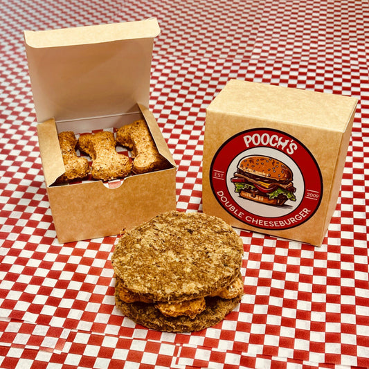A packaged dog treat resembling a double cheeseburger, with the product visible outside the box, placed on a red and white checkered surface.